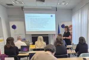 Učilnica z mizami, na katerih so prenosni računalniki, dokumenti in pisala. Več udeležencev sedi in gleda proti projekcijskemu platnu na sprednji strani prostora. Na platnu je prikazana vsebina izobraževanja z alinejami. Oseba stoji ob platnu in vodi predstavitev. Na levi strani prostora je stojalo z oglasnim panojem, na desni pa velika okna z navpičnimi žaluzijami.