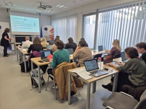 Pogled od zadaj na učilnico, kjer udeleženci sedijo za mizami z odprtimi prenosniki in gradivi. V ospredju so mize z dokumenti, stekleničkami in oblačili. Na sprednji strani prostora oseba stoji ob projekcijski površini, na kateri je prikazana predstavitev. Ob strani sta dve oglasni stojali, prostor pa osvetljujejo velika okna z navpičnimi žaluzijami.