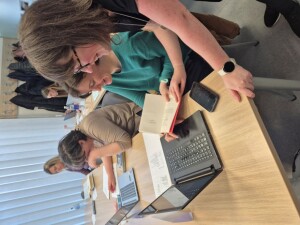 Miza z odprtim prenosnim računalnikom, dokumenti in mobilnim telefonom. Dve osebi sedita druga ob drugi, ena drži odprto knjigo, druga kaže na besedilo v njej. Na ozadju so druge osebe, ki delajo na svojih računalnikih.